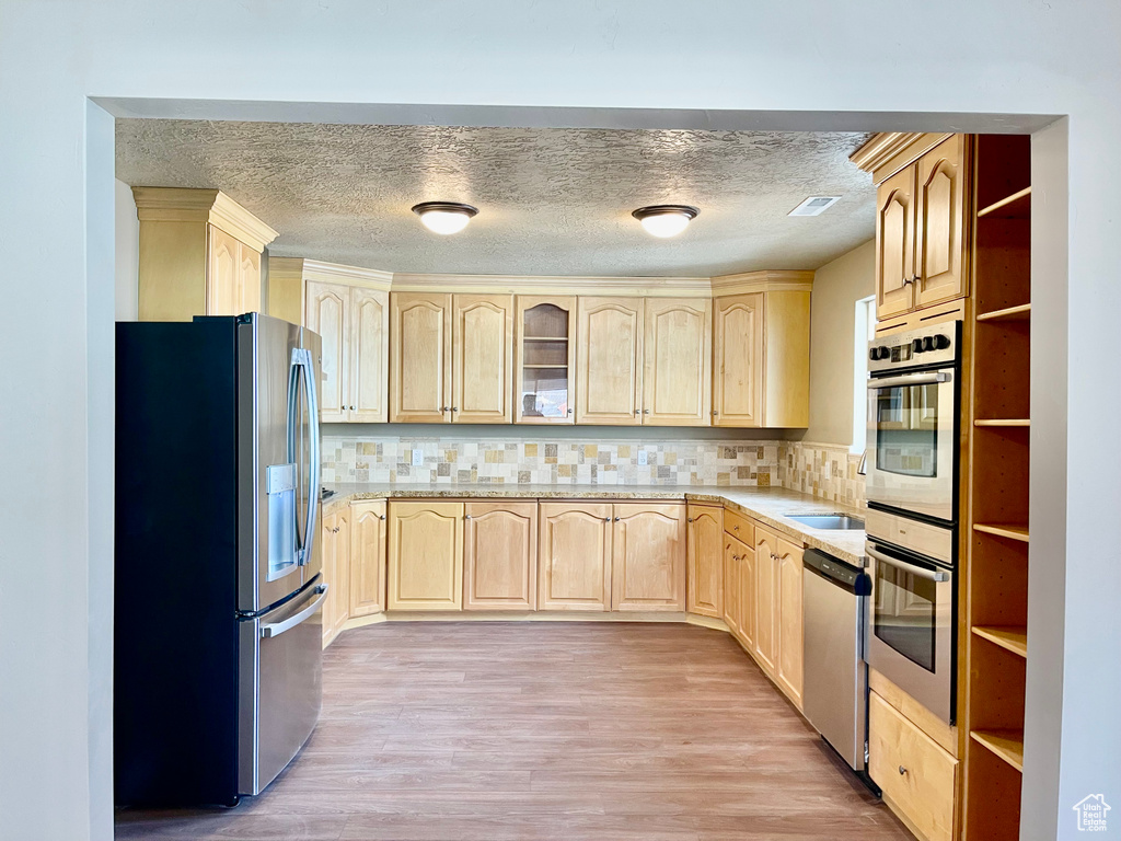 Kitchen with appliances with stainless steel finishes, light brown cabinets, a textured ceiling, decorative backsplash, and light wood-type flooring