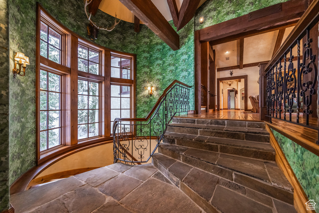 Staircase with wallpapered walls, vaulted ceiling with beams, and stone finish flooring