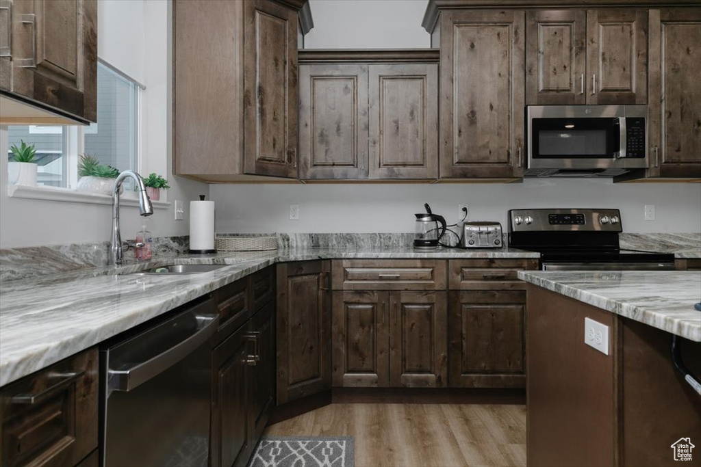 Kitchen with dark brown cabinetry, sink, light stone counters, light wood-type flooring, and appliances with stainless steel finishes