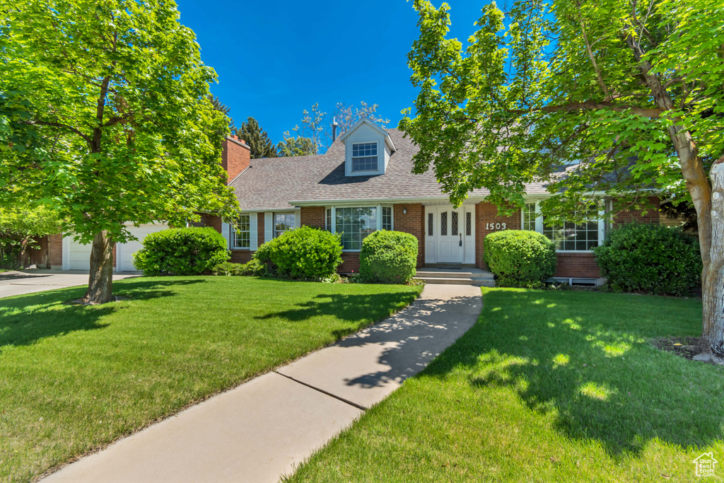 Cape cod home featuring brick siding, a front yard, roof with shingles, a garage, and a chimney