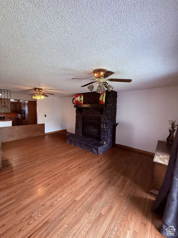 Unfurnished living room featuring ceiling fan, light wood-style floors, baseboards, a stone fireplace, and a textured ceiling