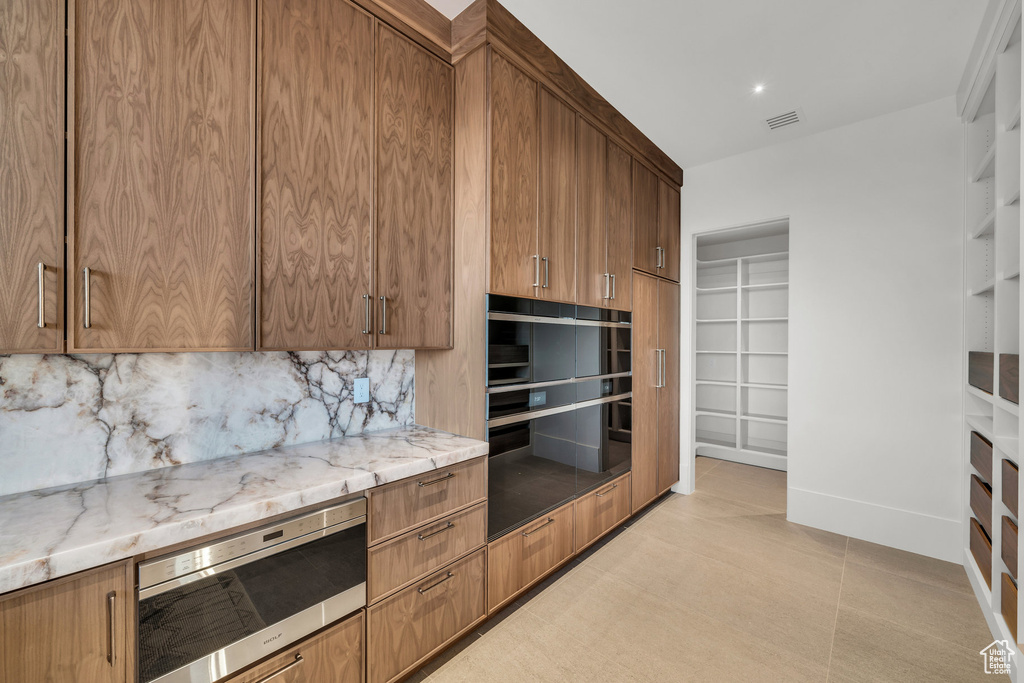Kitchen featuring stainless steel microwave, decorative backsplash, light stone counters, brown cabinets, and beverage cooler