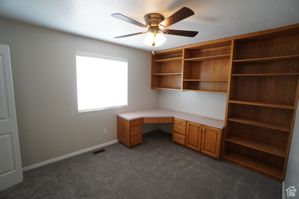 Unfurnished office with dark colored carpet, built in desk, and ceiling fan