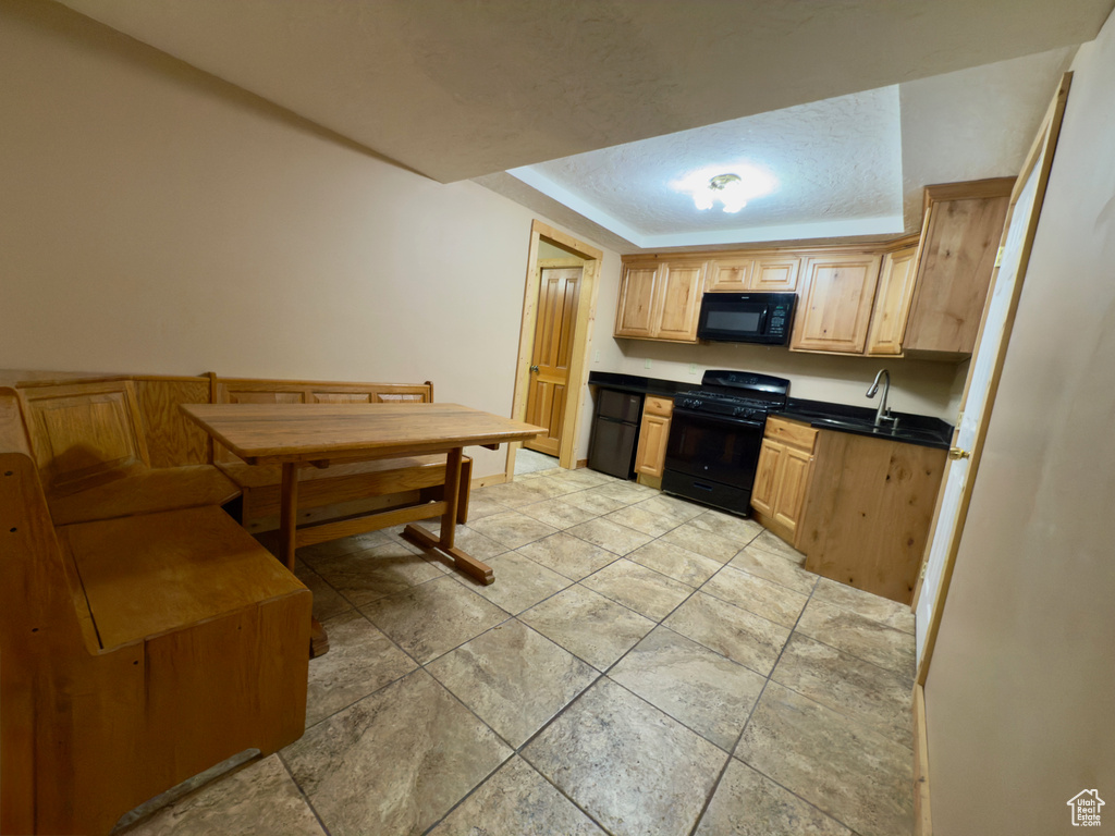 Kitchen with black appliances, dark countertops, a raised ceiling, light brown cabinetry, and a sink