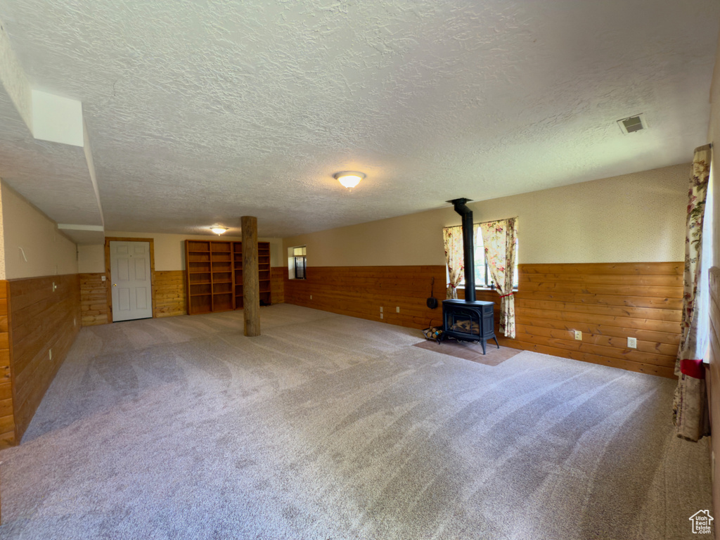 Unfurnished living room featuring wooden walls, wainscoting, a textured ceiling, a wood stove, and carpet flooring