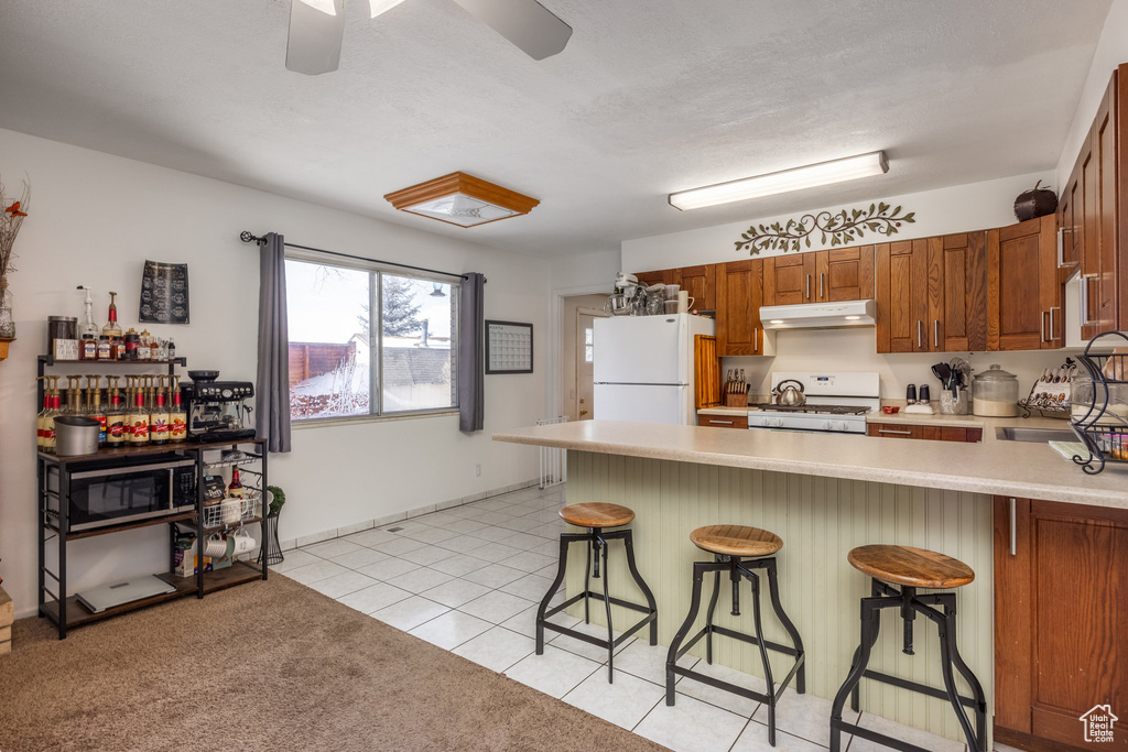 Kitchen featuring white appliances, under cabinet range hood, a peninsula, light countertops, and a kitchen breakfast bar