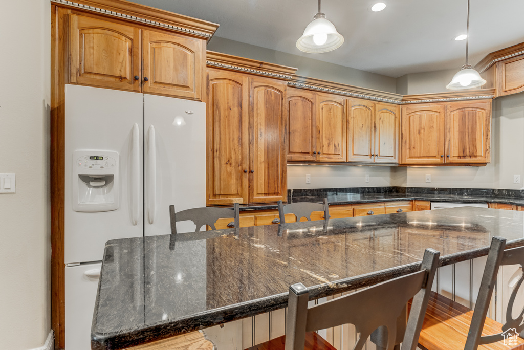 Kitchen with white fridge with ice dispenser, a kitchen bar, pendant lighting, dark stone countertops, and brown cabinetry