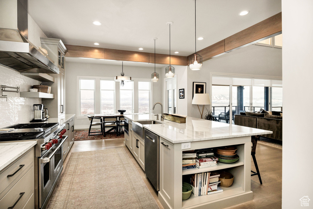 Kitchen featuring appliances with stainless steel finishes, wall chimney exhaust hood, light stone countertops, pendant lighting, and a large island with sink