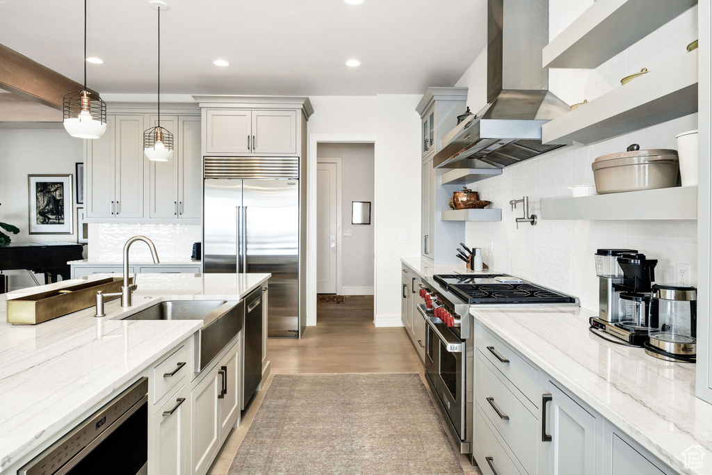 Kitchen with sink, high end appliances, wall chimney exhaust hood, hanging light fixtures, and light stone countertops