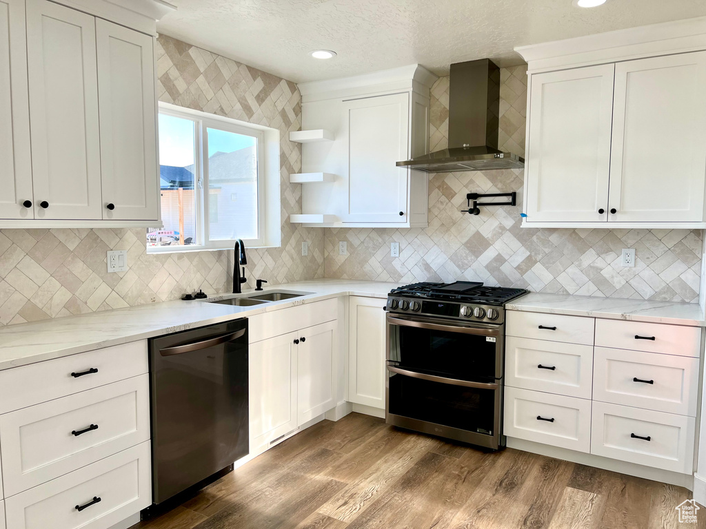 Kitchen with range with two ovens, dishwasher, wall chimney range hood, sink, and white cabinetry