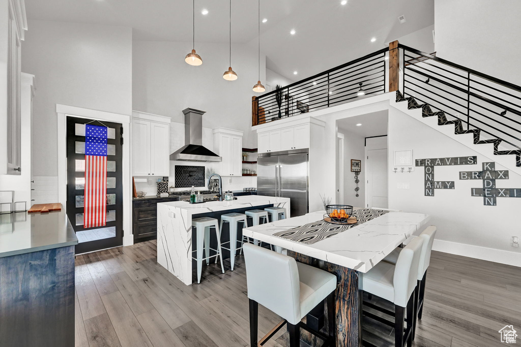 Kitchen featuring a breakfast bar, white cabinetry, a kitchen island with sink, built in fridge, and wall chimney range hood