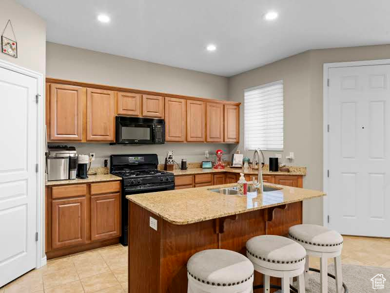 Kitchen with light tile patterned floors, sink, light stone counters, black appliances, and a kitchen island with sink