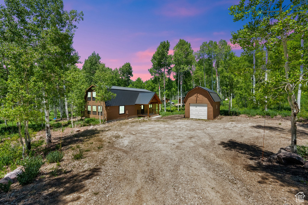 Property exterior at dusk with a detached garage, an outdoor structure, view of wooded area, and driveway