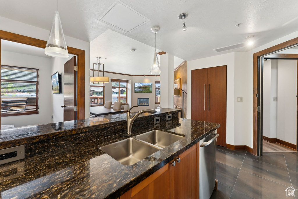 Kitchen featuring brown cabinetry, a sink, dishwasher, decorative light fixtures, and dark stone counters