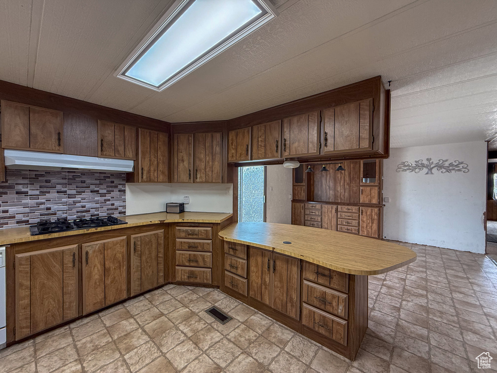 Kitchen with black gas stovetop, under cabinet range hood, a peninsula, light countertops, and decorative backsplash