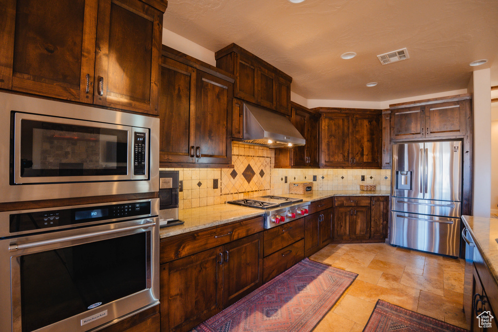 Kitchen featuring light stone countertops, wall chimney exhaust hood, appliances with stainless steel finishes, and decorative backsplash