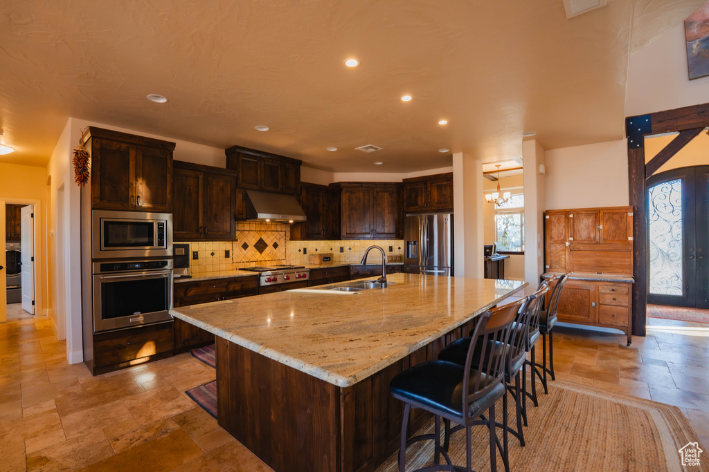 Kitchen with a sink, exhaust hood, appliances with stainless steel finishes, light stone countertops, and a large island with sink