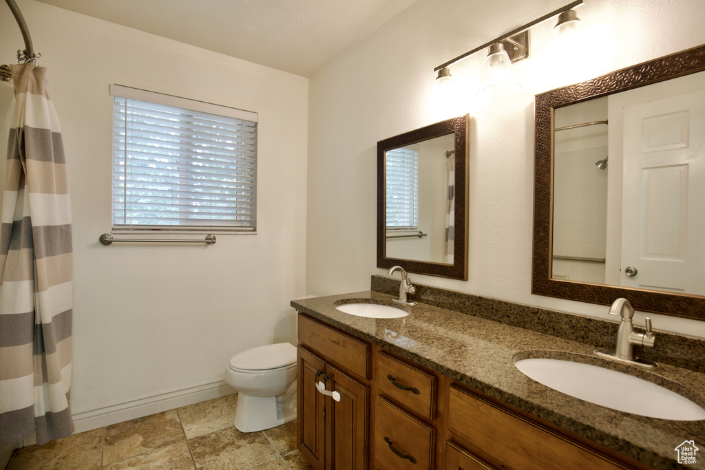 Bathroom featuring toilet, double vanity, a shower with shower curtain, and baseboards