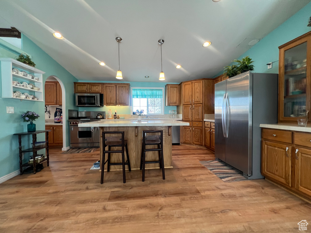 Kitchen with arched walkways, pendant lighting, stainless steel appliances, brown cabinetry, and vaulted ceiling