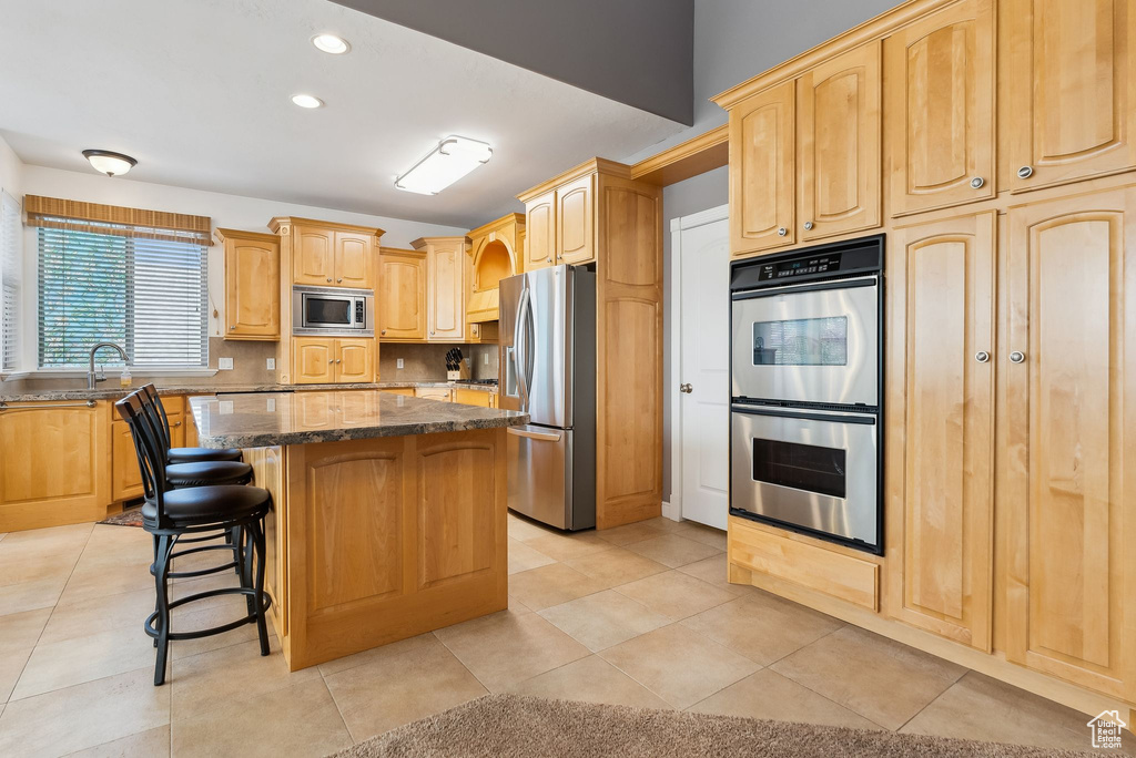Kitchen featuring backsplash, appliances with stainless steel finishes, a kitchen island, dark stone counters, and a kitchen bar
