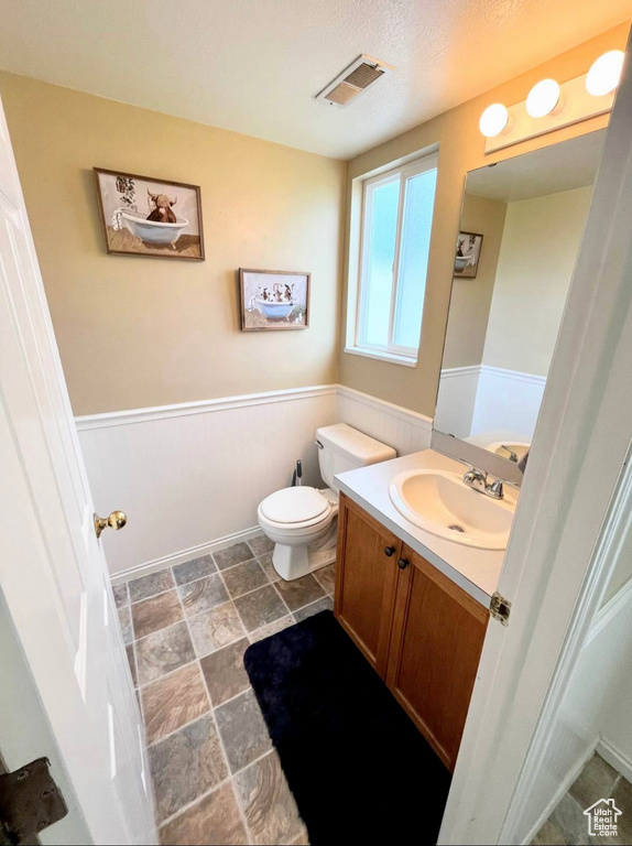 Bathroom featuring visible vents, toilet, stone finish floor, wainscoting, and vanity