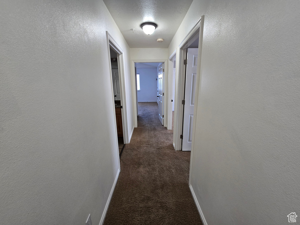 Hall featuring a textured wall, baseboards, and dark carpet