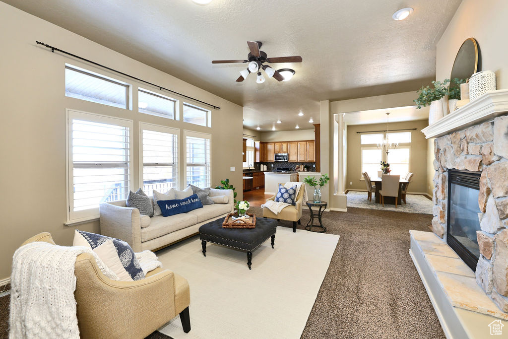 Living area featuring baseboards, light colored carpet, a textured ceiling, a stone fireplace, and ceiling fan with notable chandelier
