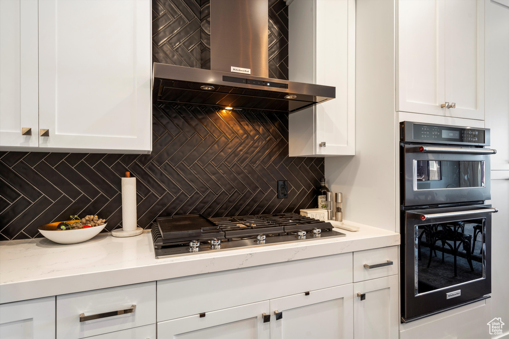 Kitchen with double oven, white cabinets, stainless steel gas cooktop, and wall chimney range hood
