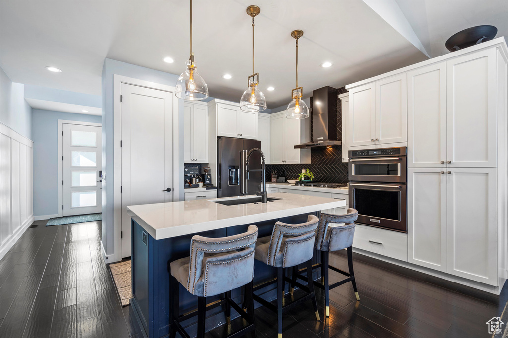 Kitchen with a kitchen island with sink, white cabinets, hanging light fixtures, wall chimney range hood, and light countertops