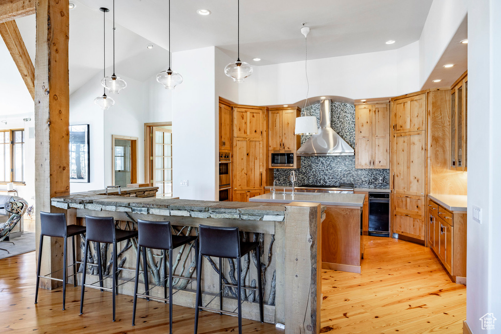 Kitchen featuring wall chimney range hood, light wood finished floors, decorative backsplash, stainless steel appliances, and a breakfast bar area