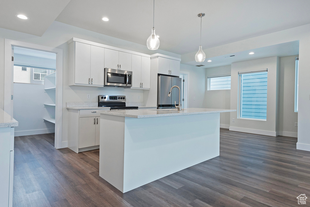 Kitchen featuring dark wood-style flooring, stainless steel appliances, hanging light fixtures, white cabinetry, and an island with sink