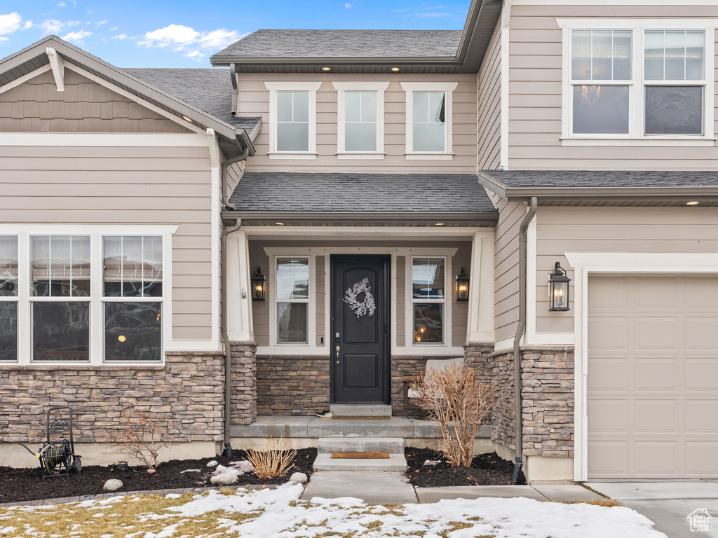 Snow covered property entrance with a garage, stone siding, and roof with shingles