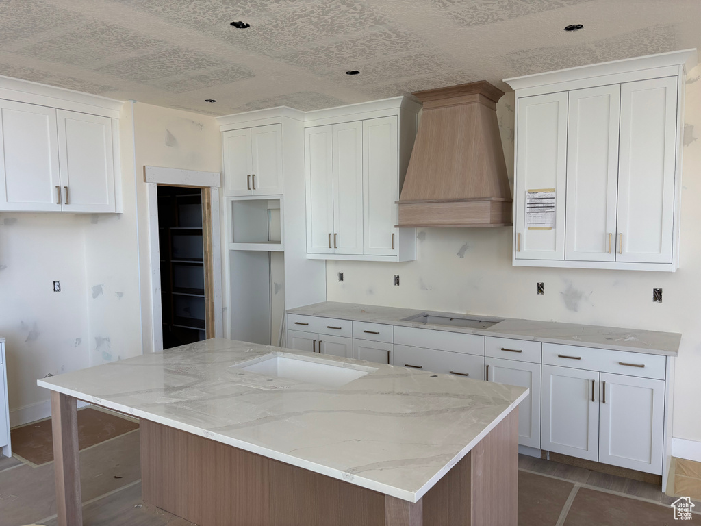 Kitchen with black stovetop, premium range hood, light stone countertops, a kitchen island, and white cabinetry