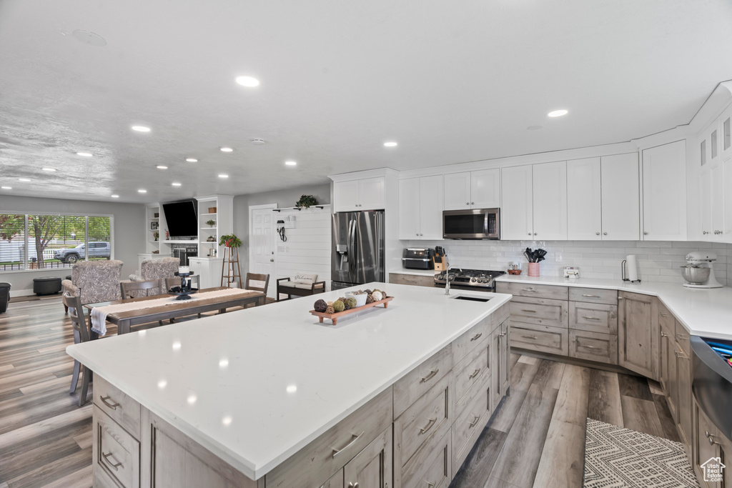 Kitchen featuring stainless steel appliances, wood finished floors, recessed lighting, a center island with sink, and light countertops