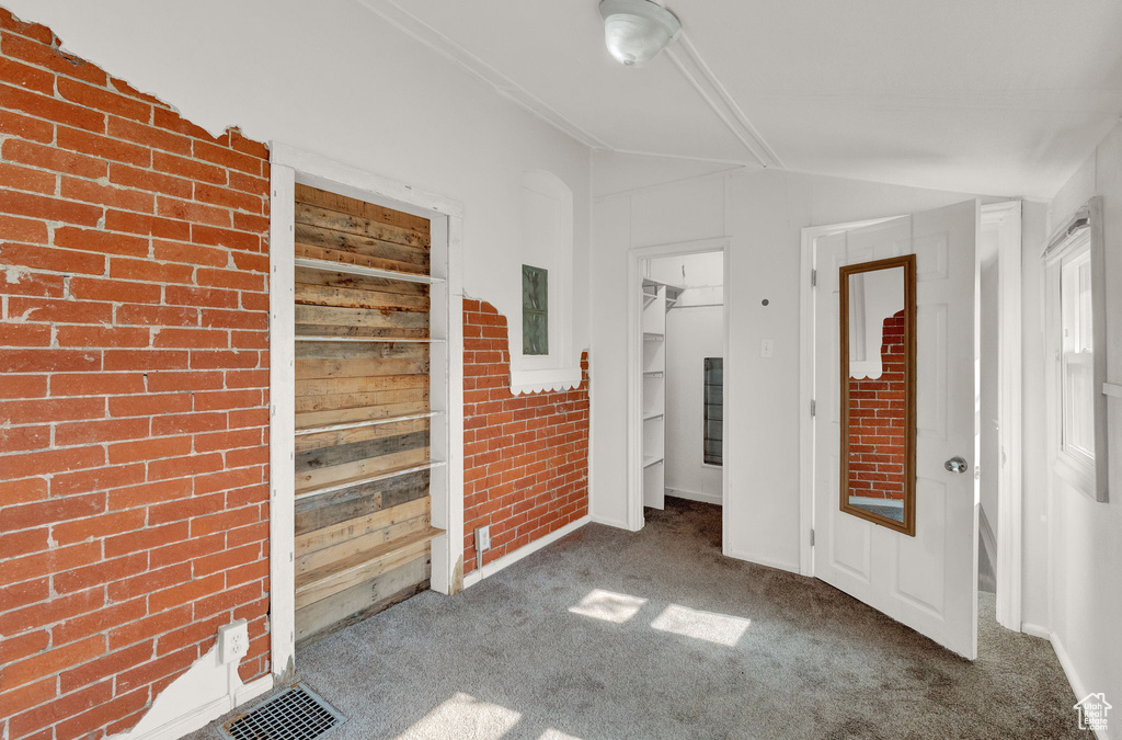 Unfurnished bedroom featuring dark colored carpet, visible vents, a spacious closet, vaulted ceiling, and brick wall