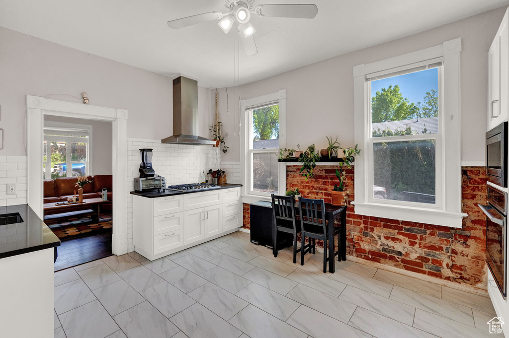 Kitchen with dark countertops, ceiling fan, wall chimney exhaust hood, stainless steel appliances, and white cabinetry