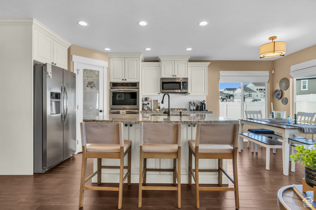 Kitchen with dark wood-style flooring, recessed lighting, appliances with stainless steel finishes, a kitchen island with sink, and light stone countertops