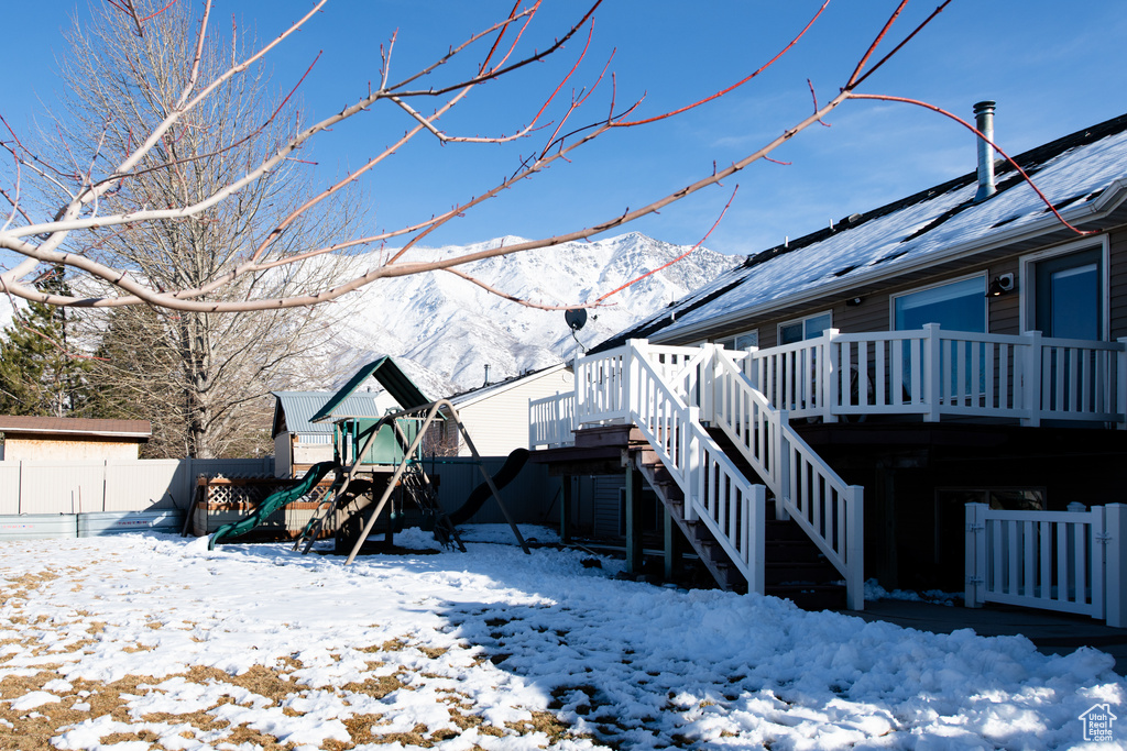 Snow covered rear of property with a playground, fence, a deck with mountain view, and stairs