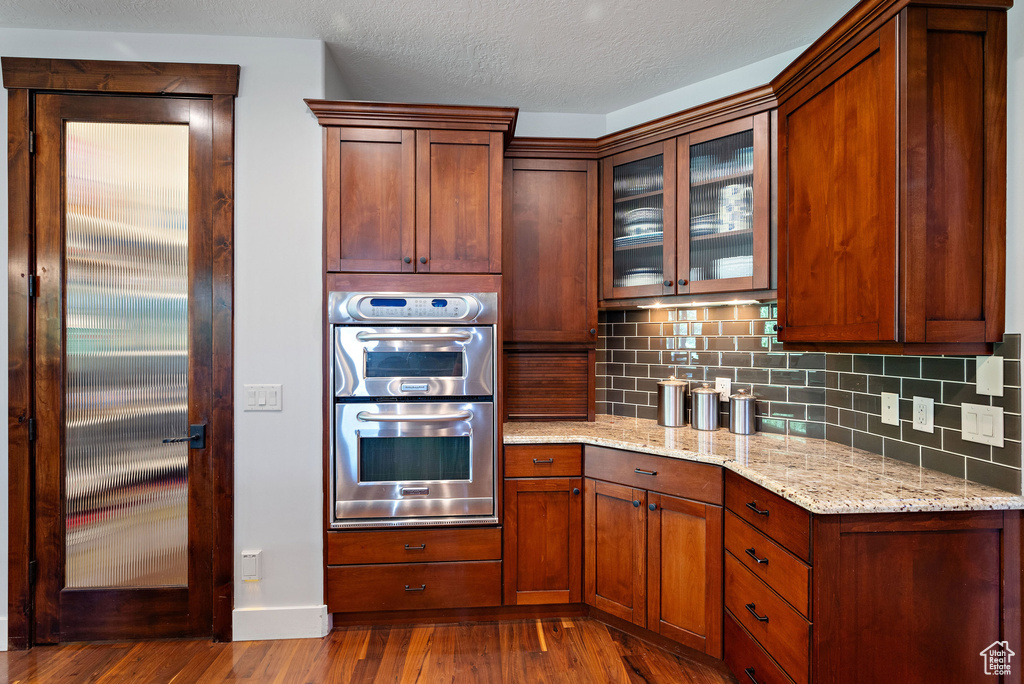 Kitchen with stainless steel double oven, light stone counters, backsplash, dark wood-style floors, and glass insert cabinets