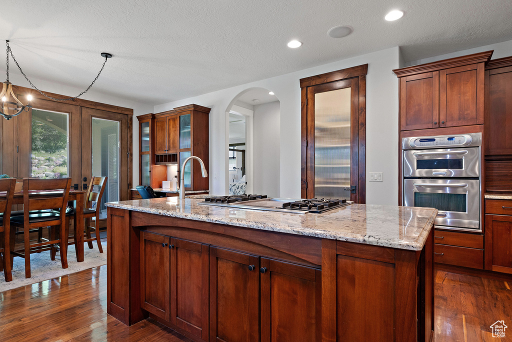 Kitchen with appliances with stainless steel finishes, arched walkways, light stone countertops, dark wood-type flooring, and an island with sink