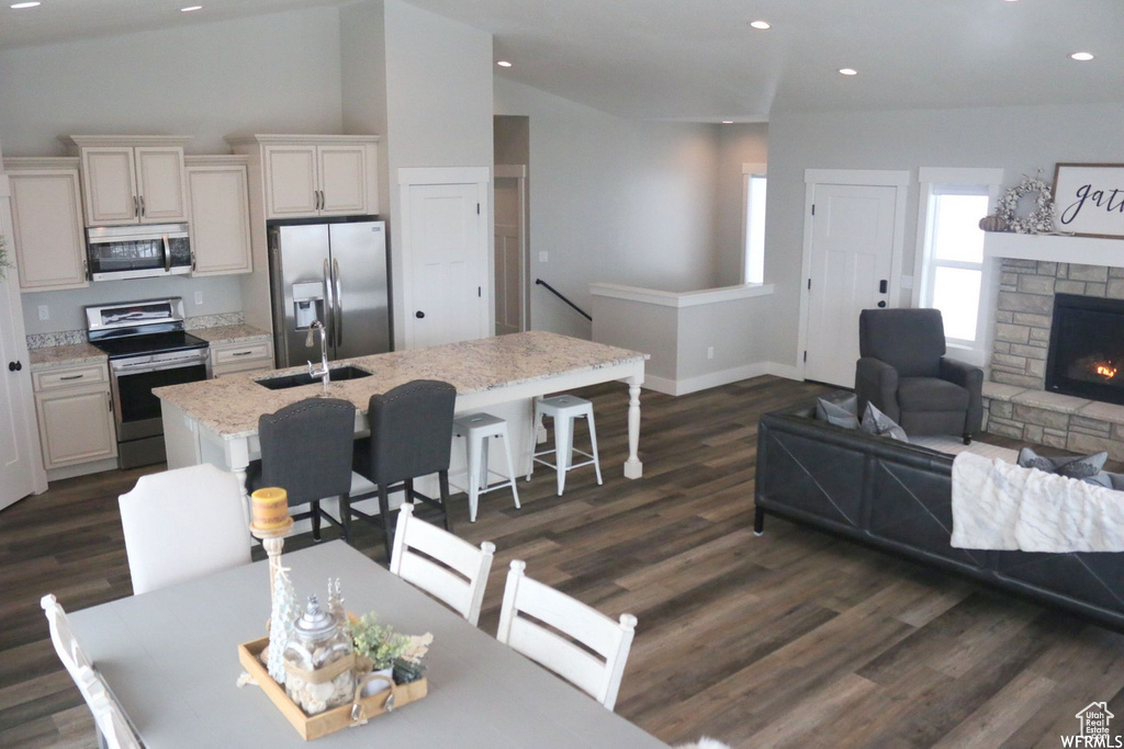 Kitchen featuring a stone fireplace, dark wood finished floors, appliances with stainless steel finishes, an island with sink, and a kitchen bar