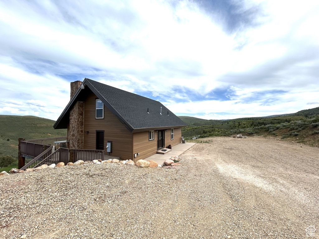 View of side of home featuring a chimney, driveway, roof with shingles, a wooden deck, and a patio area