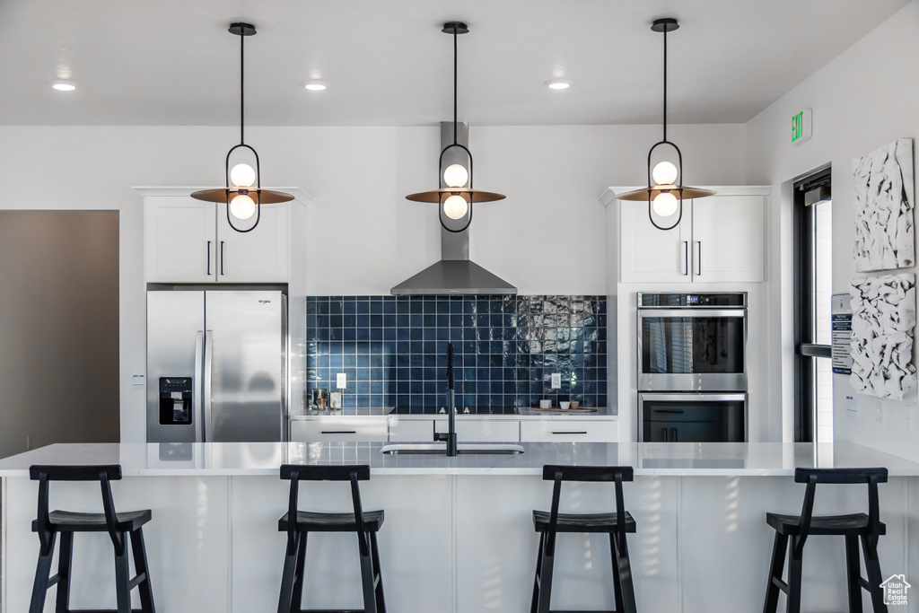 Kitchen featuring white cabinets, wall chimney exhaust hood, stainless steel appliances, pendant lighting, and backsplash
