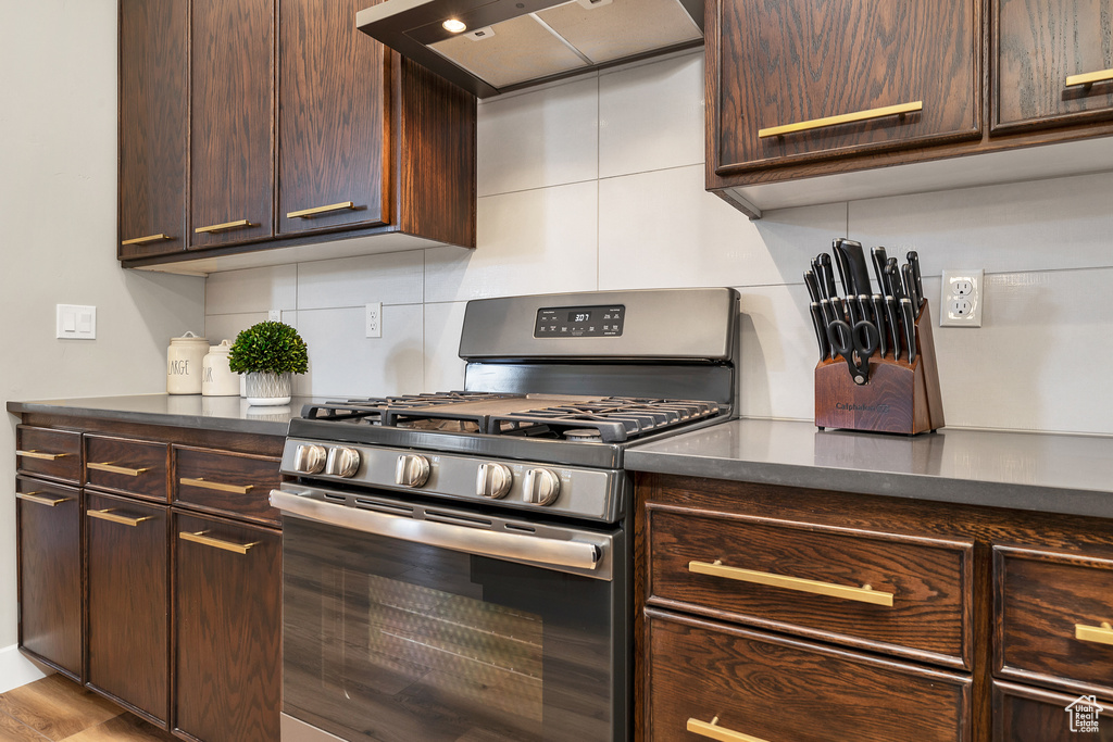Kitchen featuring stainless steel gas range, tasteful backsplash, dark brown cabinetry, wall chimney range hood, and dark countertops