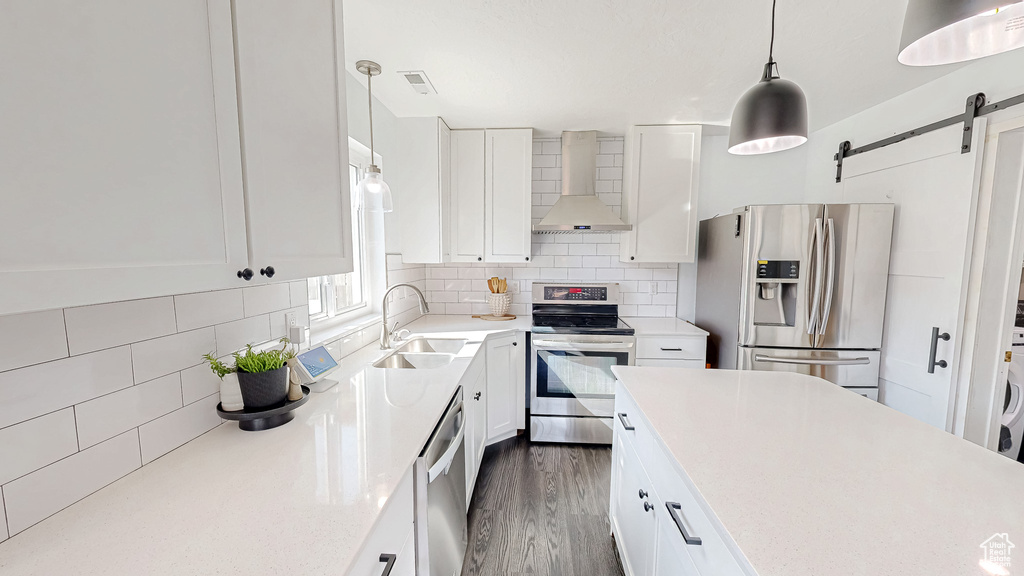 Kitchen with a barn door, decorative backsplash, wall chimney exhaust hood, appliances with stainless steel finishes, and a sink