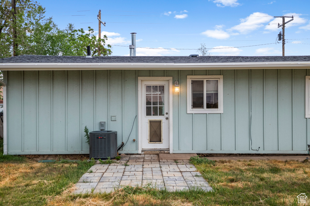 Doorway to property featuring board and batten siding, cooling unit, roof with shingles, and a yard