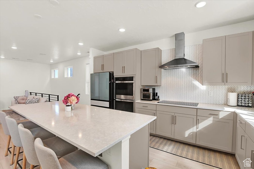 Kitchen featuring double oven, black electric stovetop, gray cabinetry, wall chimney range hood, and freestanding refrigerator