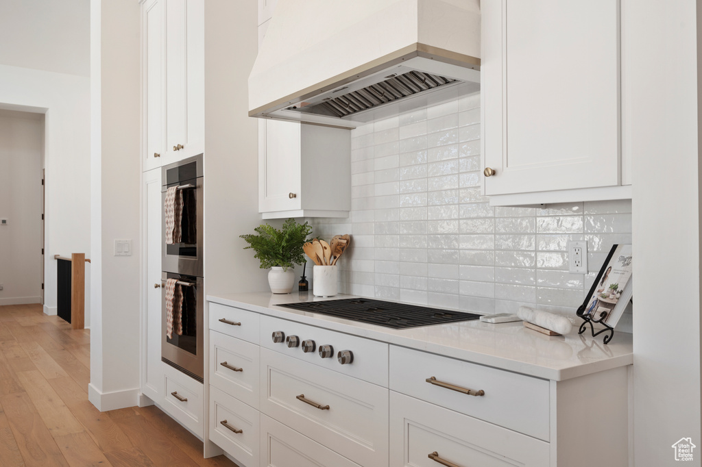 Kitchen featuring black cooktop, tasteful backsplash, custom exhaust hood, double oven, and white cabinetry