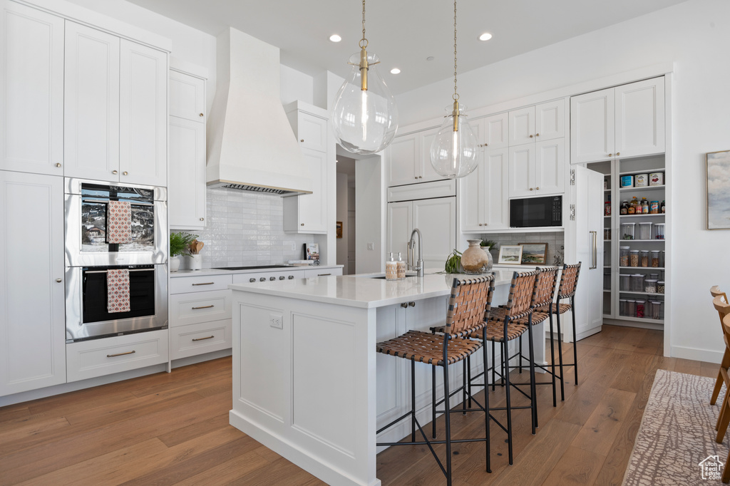 Kitchen with light countertops, light wood-style floors, white cabinets, built in appliances, and premium range hood