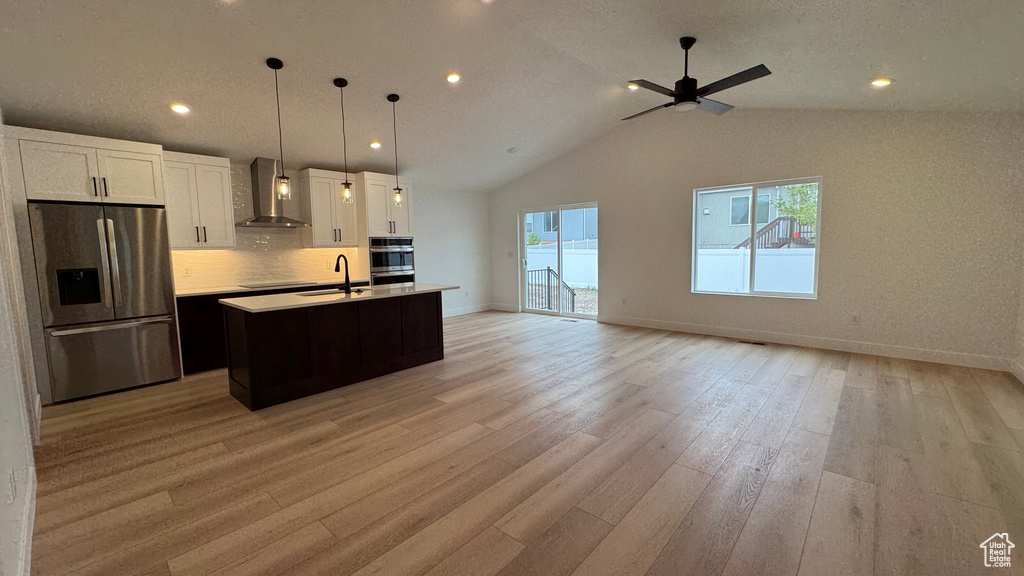 Kitchen featuring wall chimney range hood, a kitchen island with sink, appliances with stainless steel finishes, a sink, and open floor plan